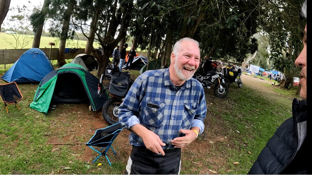 Man in a checkered shirt standing in a camping area with tents and people around.